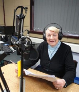 Joy is sitting alone in the Talking Newspaper studio, smiling at the camera. A small stack of recording equipment is behind her, in the corner of the room. Joy is wearing a black pair of headphones, and is in front of one of the microphones; resting her hands on the table, holding a newspaper - which she is preparing to read from.